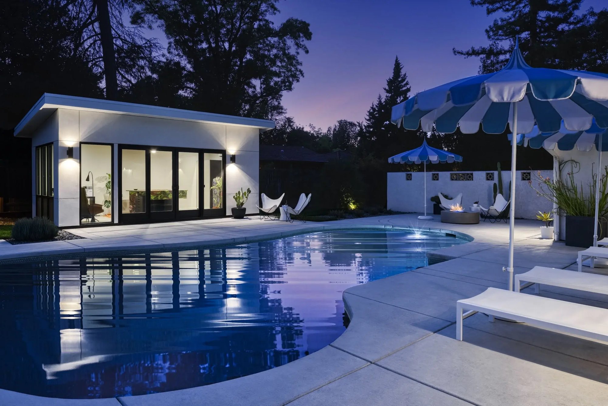 Modern pool house at dusk with illuminated patio, pool, lounge chairs, and striped umbrellas. Modern pool house at dusk with illuminated patio, pool, lounge chairs, and striped umbrellas.